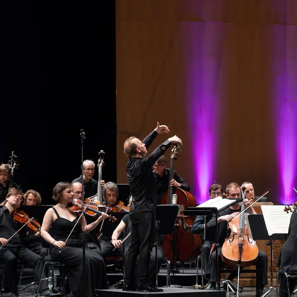 Ein Orchester spielt auf der Bühne, der Dirigent steht mit gehobenen Armen vor den Musikern.