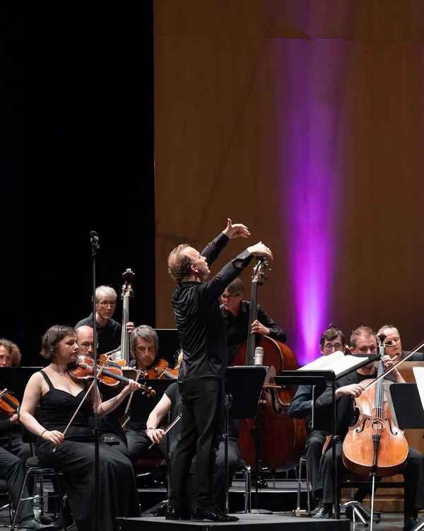 Ein Orchester spielt auf der Bühne, der Dirigent steht mit gehobenen Armen vor den Musikern.