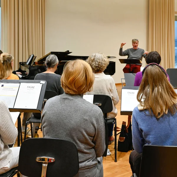 Mehrere Personen sitzen bei einer Chorprobe mit dem Rücken zur Kamera und schauen auf den Chorleiter.