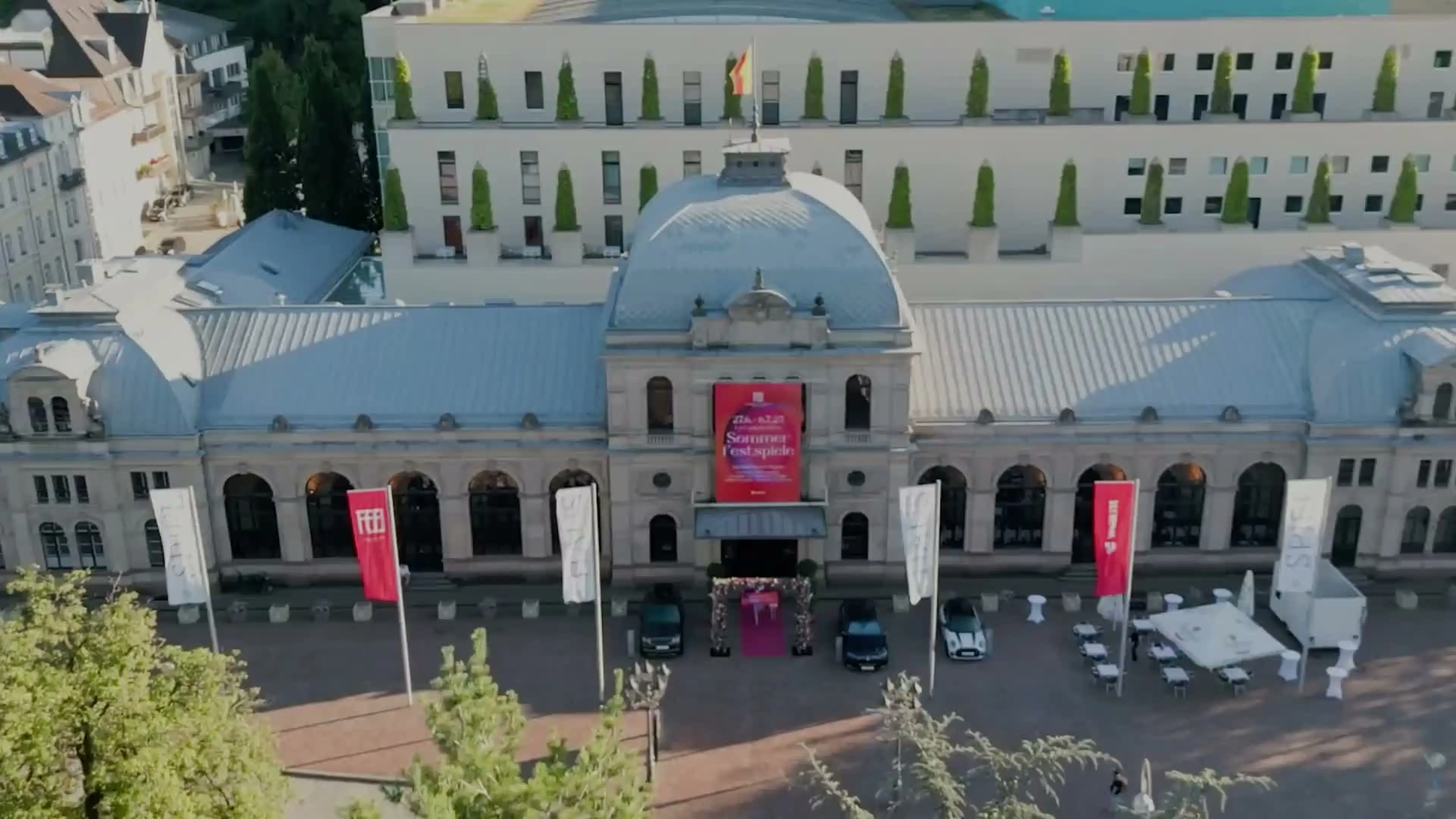 Luftaufnahme eines historischen Festspielhaus Gebäudes mit gewölbtem Dach und roten Bannern. Es befindet sich umgeben von Bäumen und Architektur.