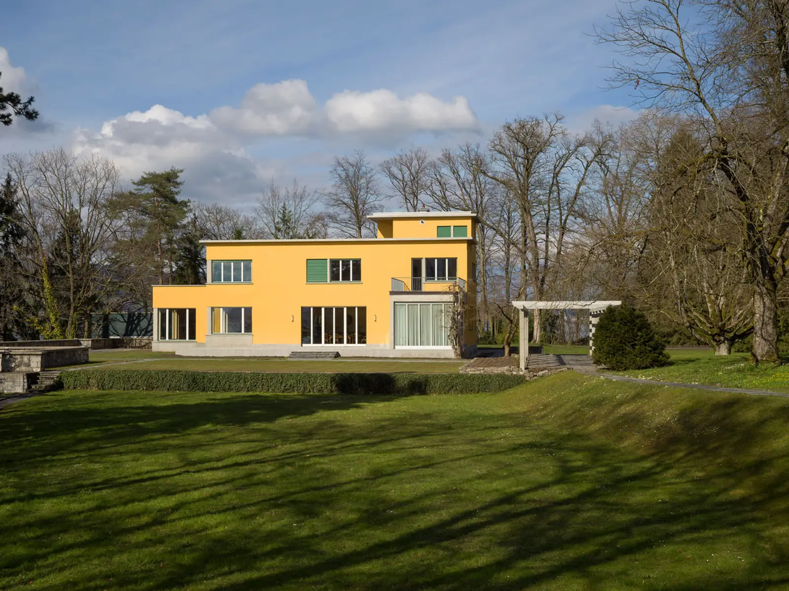 Sergei Rachmaninows Villa am Vierwaldstätter See. Ein gelb angestrichenes Haus inmitten von grünem Rasen, Bäumen und blauem Himmel