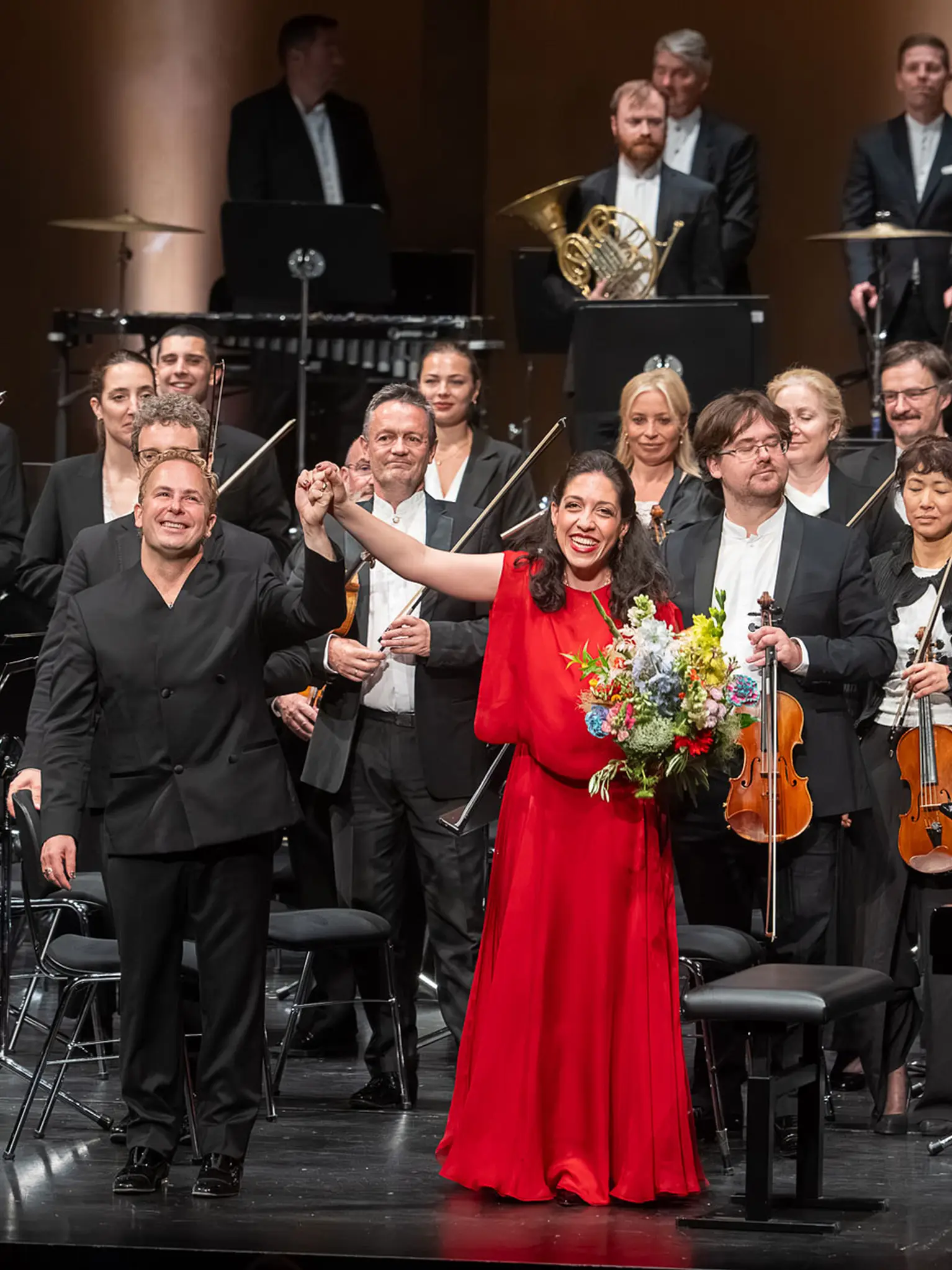 Orchester, Dirigent und Klaviersolistin stehen auf einer Bühne beim Schlussapplaus. Die Pianistin trägt ein rotes Kleid und hält einen Blumenstrauß.