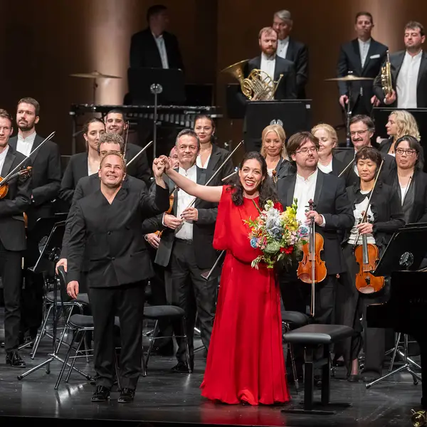 Orchester, Dirigent und Klaviersolistin stehen auf einer Bühne beim Schlussapplaus. Die Pianistin trägt ein rotes Kleid und hält einen Blumenstrauß.