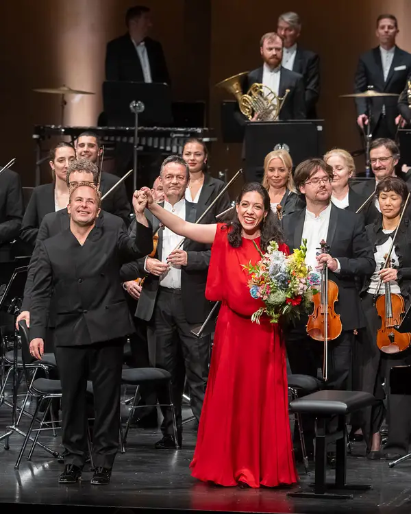 Orchester, Dirigent und Klaviersolistin stehen auf einer Bühne beim Schlussapplaus. Die Pianistin trägt ein rotes Kleid und hält einen Blumenstrauß.