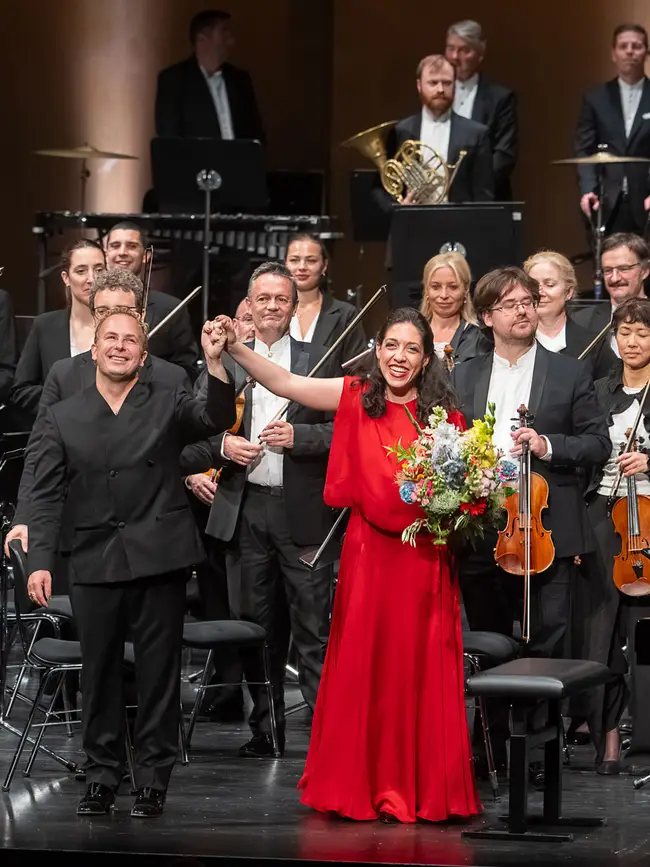 Orchester, Dirigent und Klaviersolistin stehen auf einer Bühne beim Schlussapplaus. Die Pianistin trägt ein rotes Kleid und hält einen Blumenstrauß.