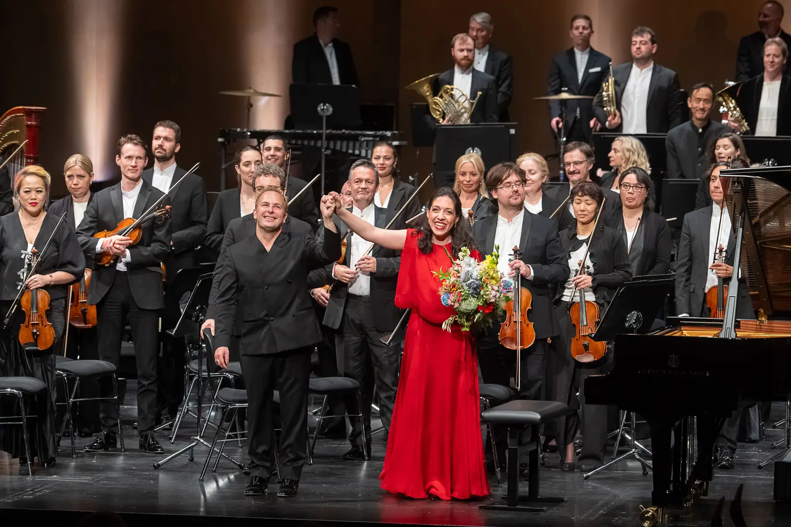 Orchester, Dirigent und Klaviersolistin stehen auf einer Bühne beim Schlussapplaus. Die Pianistin trägt ein rotes Kleid und hält einen Blumenstrauß.