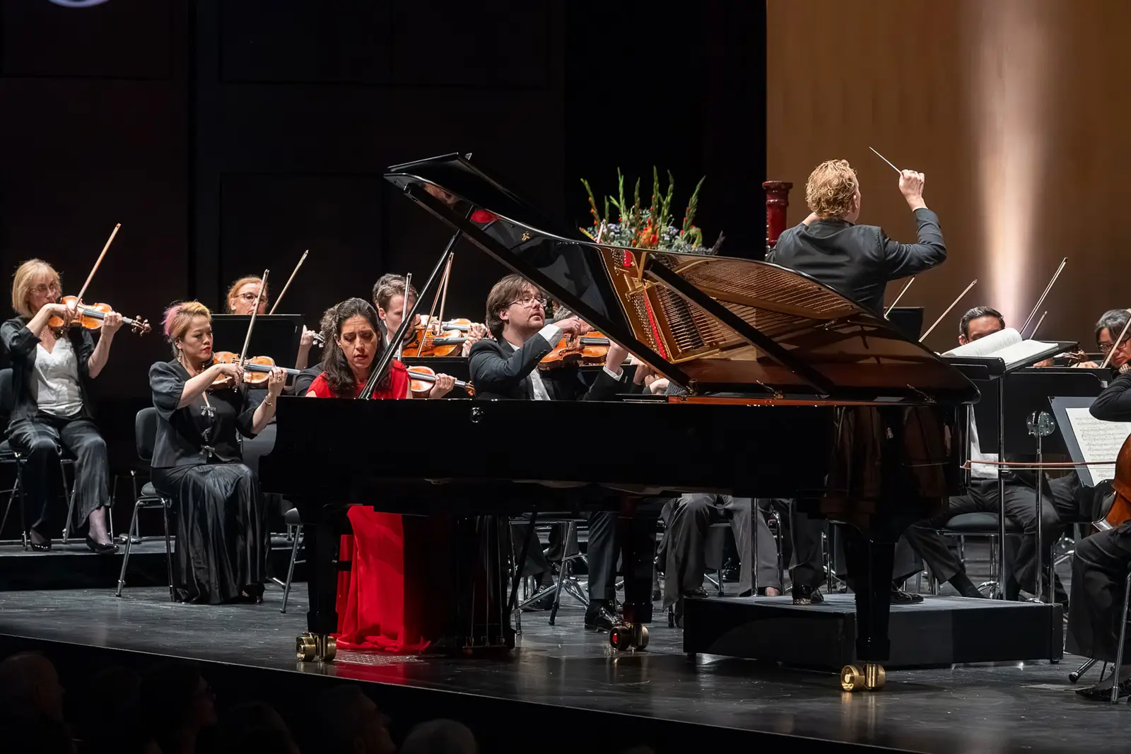Ein Orchester spielt auf einer großen Bühne. Am Klavier sitzt eine Pianistin in rotem Kleid, der Dirigent steht vor dem Orchester.