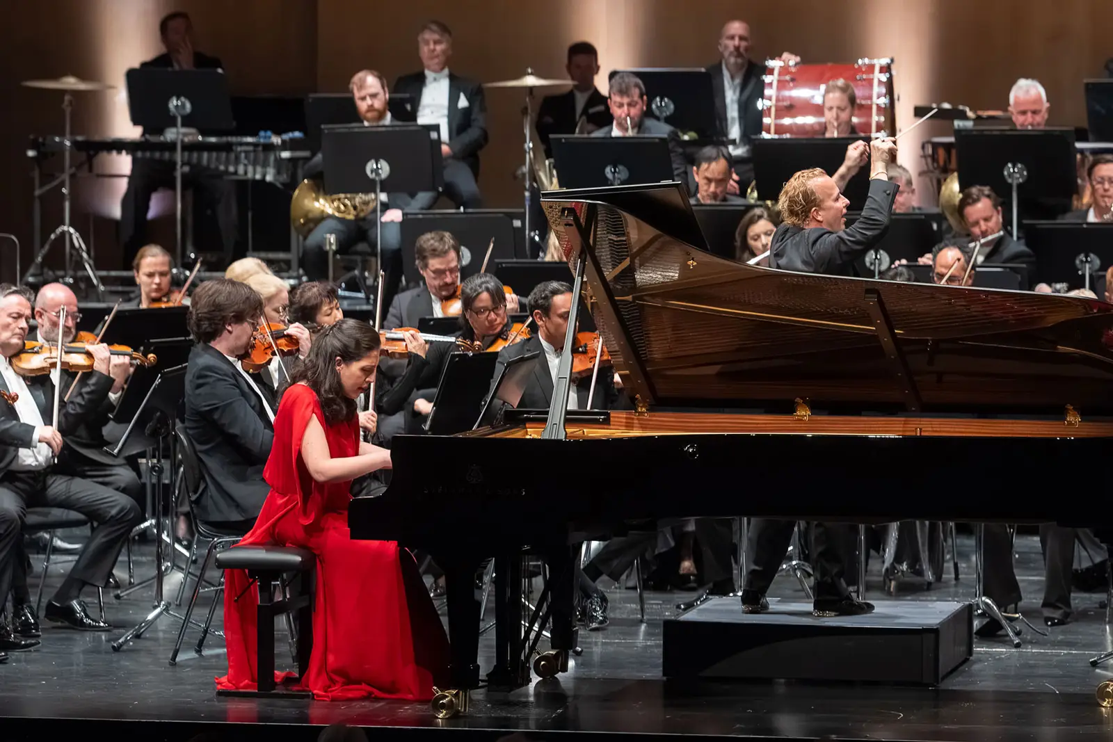 Eine Pianistin in rotem Kleid sitzt am Flügel und spielt. Hinter ihr sind ein Dirigent und ein Orchester zu sehen.