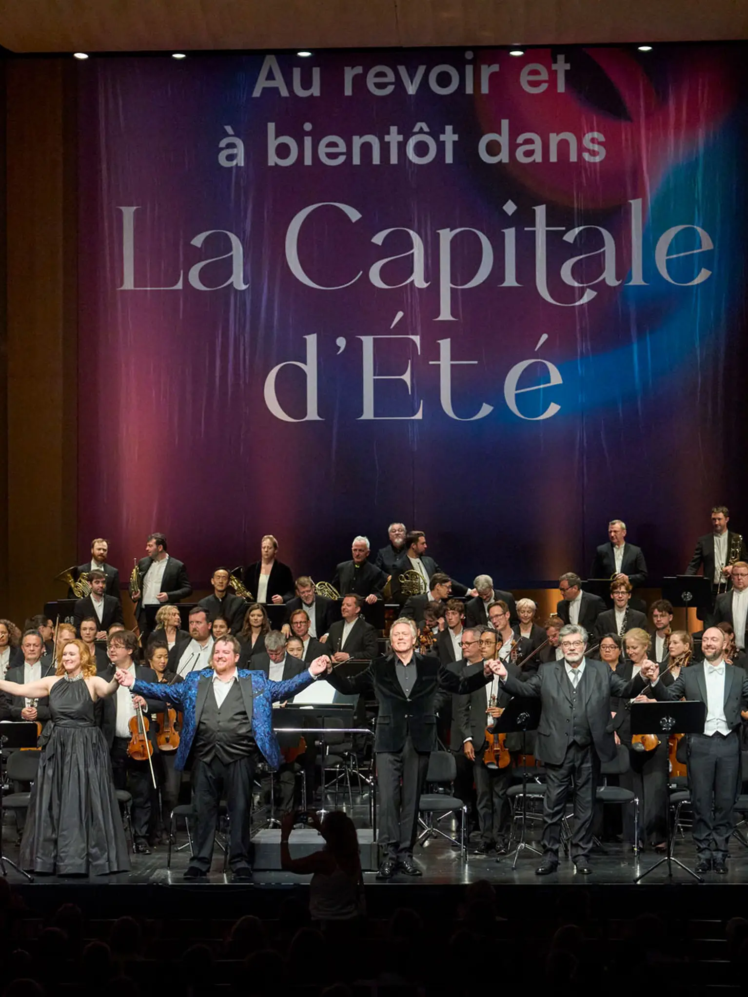 Blick auf eine große Bühne. Vor dem Orchester stehen neun Personen und verbeugen sich. Im Hintergrund ist ein Transparent mit der Aufschrift "Au revoir et à bientôt dans La Capitale d'Été" zu sehen.