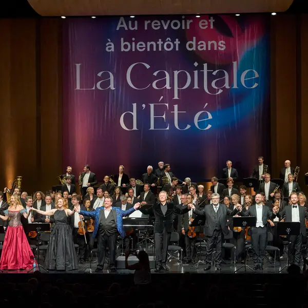 Blick auf eine große Bühne. Vor dem Orchester stehen neun Personen und verbeugen sich. Im Hintergrund ist ein Transparent mit der Aufschrift "Au revoir et à bientôt dans La Capitale d'Été" zu sehen.