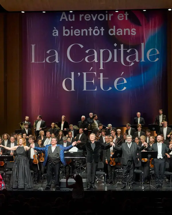 Blick auf eine große Bühne. Vor dem Orchester stehen neun Personen und verbeugen sich. Im Hintergrund ist ein Transparent mit der Aufschrift "Au revoir et à bientôt dans La Capitale d'Été" zu sehen.