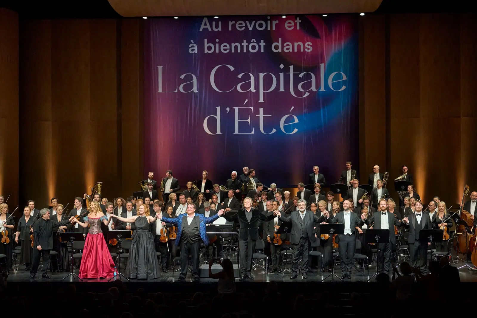 Blick auf eine große Bühne. Vor dem Orchester stehen neun Personen und verbeugen sich. Im Hintergrund ist ein Transparent mit der Aufschrift "Au revoir et à bientôt dans La Capitale d'Été" zu sehen.
