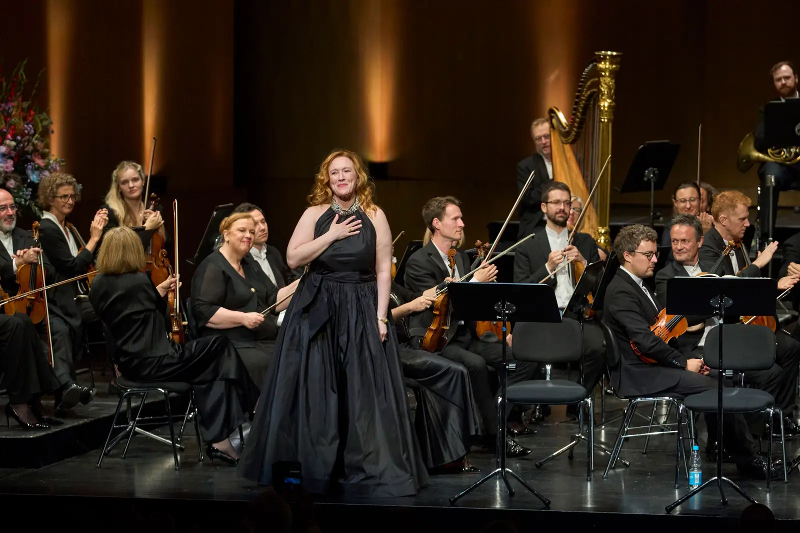 Eine Sängerin in schwarzem Kleid steht auf einer Bühne vor dem Orchester und lächelt ins Publikum, ihre Hand liegt auf ihrem Herzen.