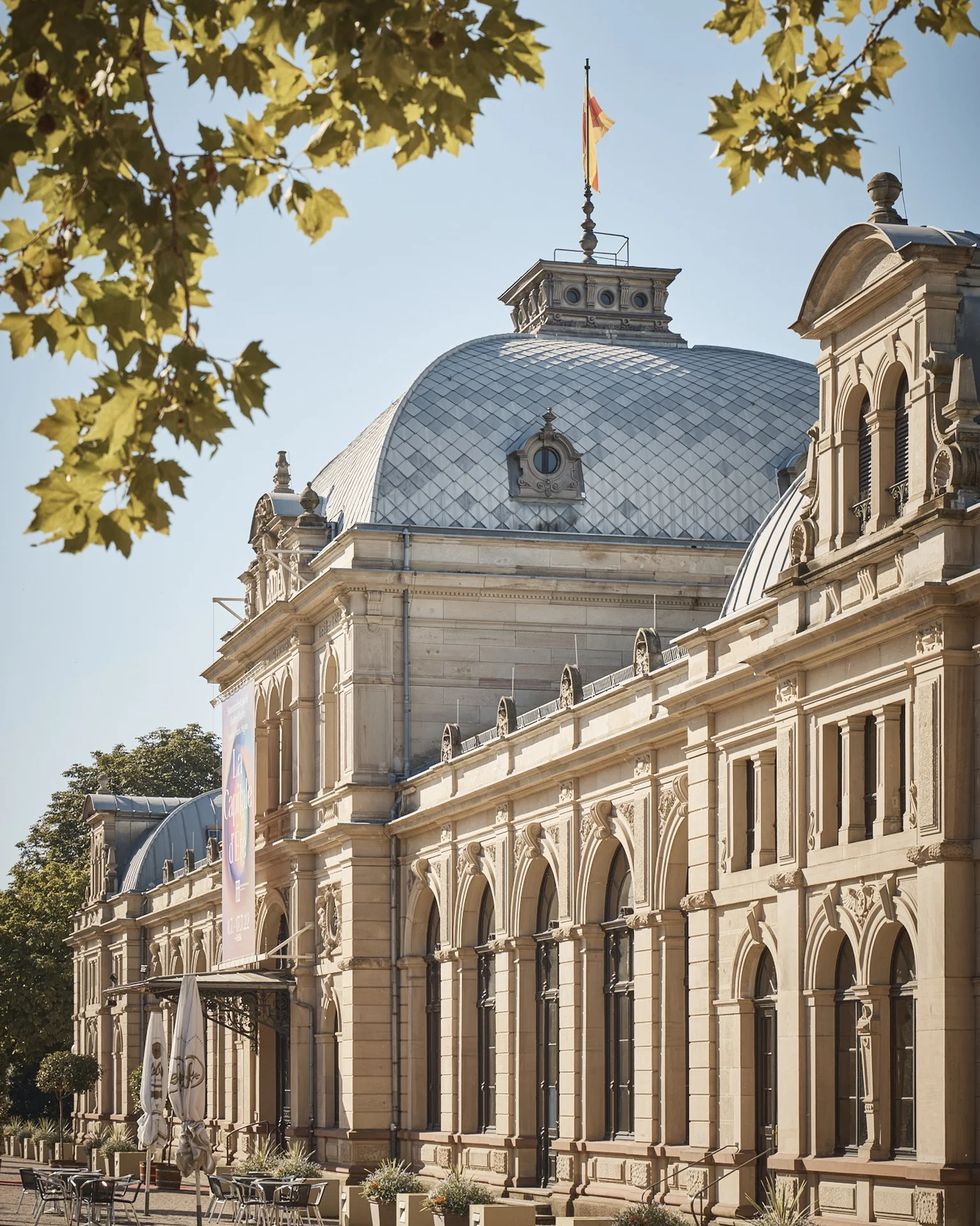Das Festspielhaus im klassischen Stil mit gewölbtem Dach, flankiert von Bäumen. Eine Fahne weht auf der Kuppel.