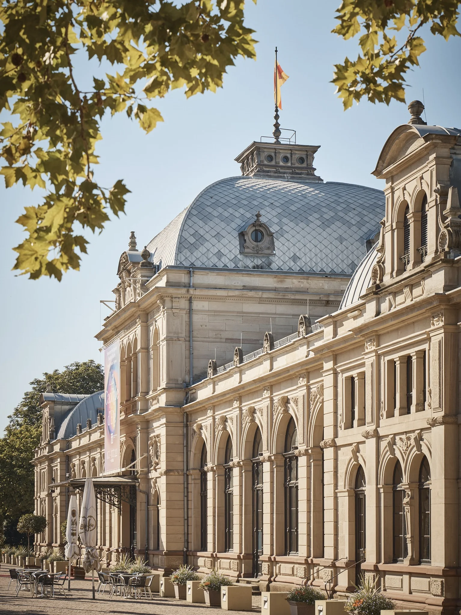 Das Festspielhaus im klassischen Stil mit gewölbtem Dach, flankiert von Bäumen. Eine Fahne weht auf der Kuppel.