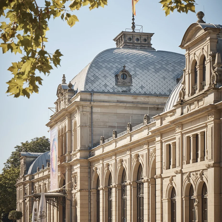 Das Festspielhaus im klassischen Stil mit gewölbtem Dach, flankiert von Bäumen. Eine Fahne weht auf der Kuppel.