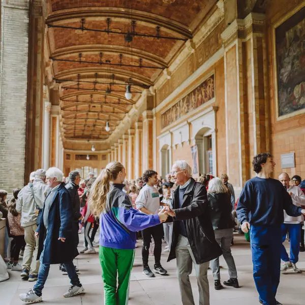 Des personnes dansent et interagissent dans une grande salle historique, avec des plafonds voûtés et des murs décorés, créant une ambiance animée.