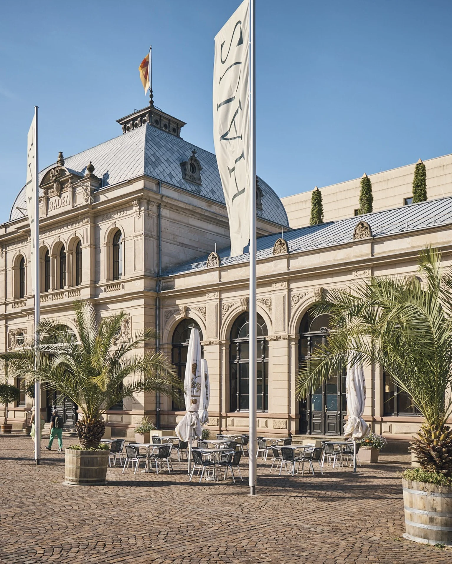 Das Bild zeigt ein elegantes, historisches Gebäude mit Palmen und Fahnen davor, unter blauem Himmel.