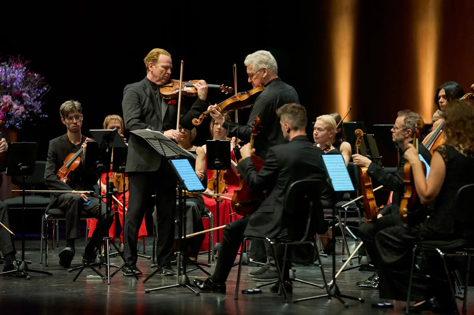 Zwei Violinisten stehen sich gegenüber und machen Musik, um sie herum sitzt ein Orchester.