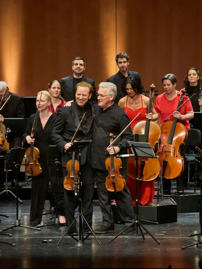 Ein Orchester steht auf einer großen Bühne. Im Vordergrund stehen 2 Violinisten die beim Applaus freundlich ins Publikum blicken.