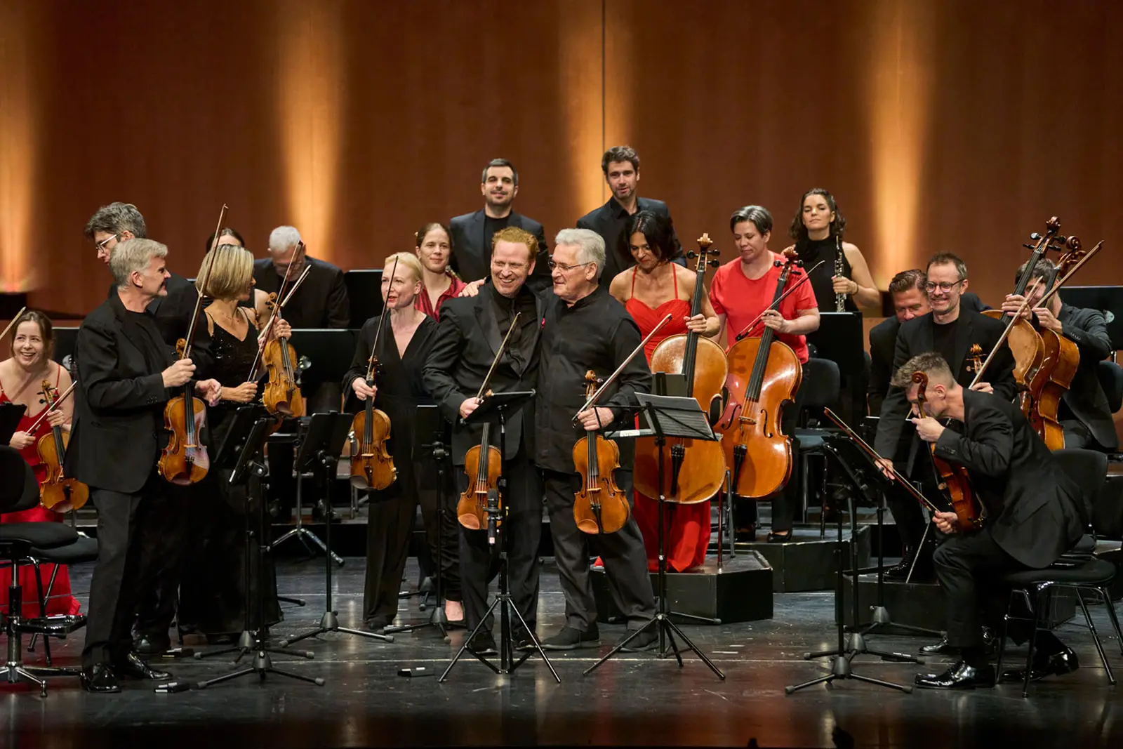 Ein Orchester steht auf einer großen Bühne. Im Vordergrund stehen 2 Violinisten die beim Applaus freundlich ins Publikum blicken.