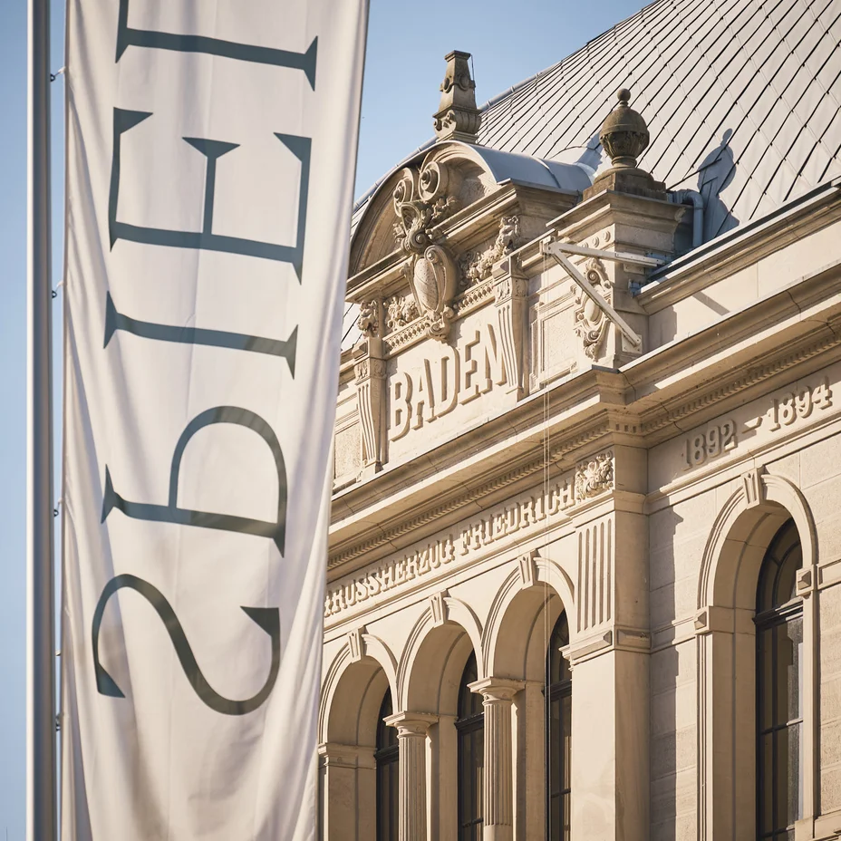 Außenansicht eines historischen Gebäudes mit Aufschrift "Baden". Ein großes Banner zeigt das Wort "SPIELT" in stilvoller Schrift.