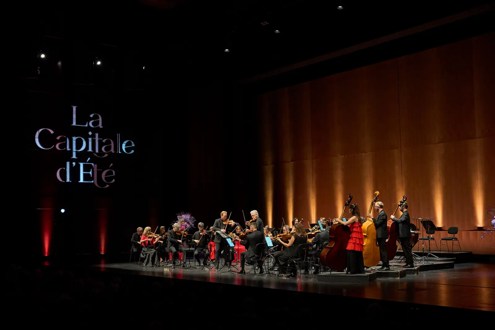Blick aus dem Konzertsaal auf ein Orchester auf der Bühne. Der Hintergrund ist orange beleuchtet und auf die linke Seite der Bühne wird der Schriftzug "La Capitale d'Été" projiziert.