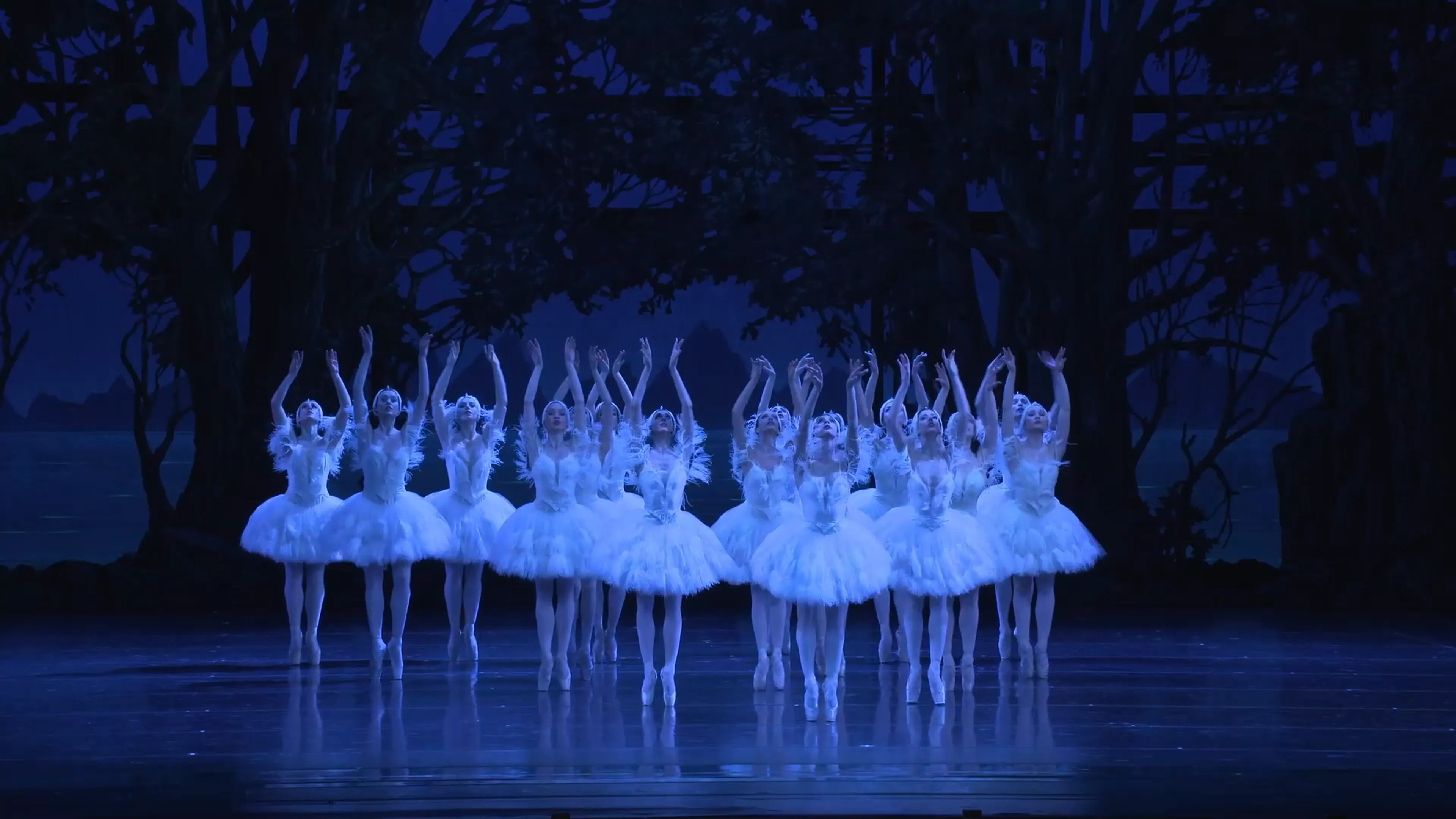 Eine Gruppe Balletttänzerinnen in weißer Kleidung tanzt auf einer Bühne. Die Szene ist blau beleuchtet.