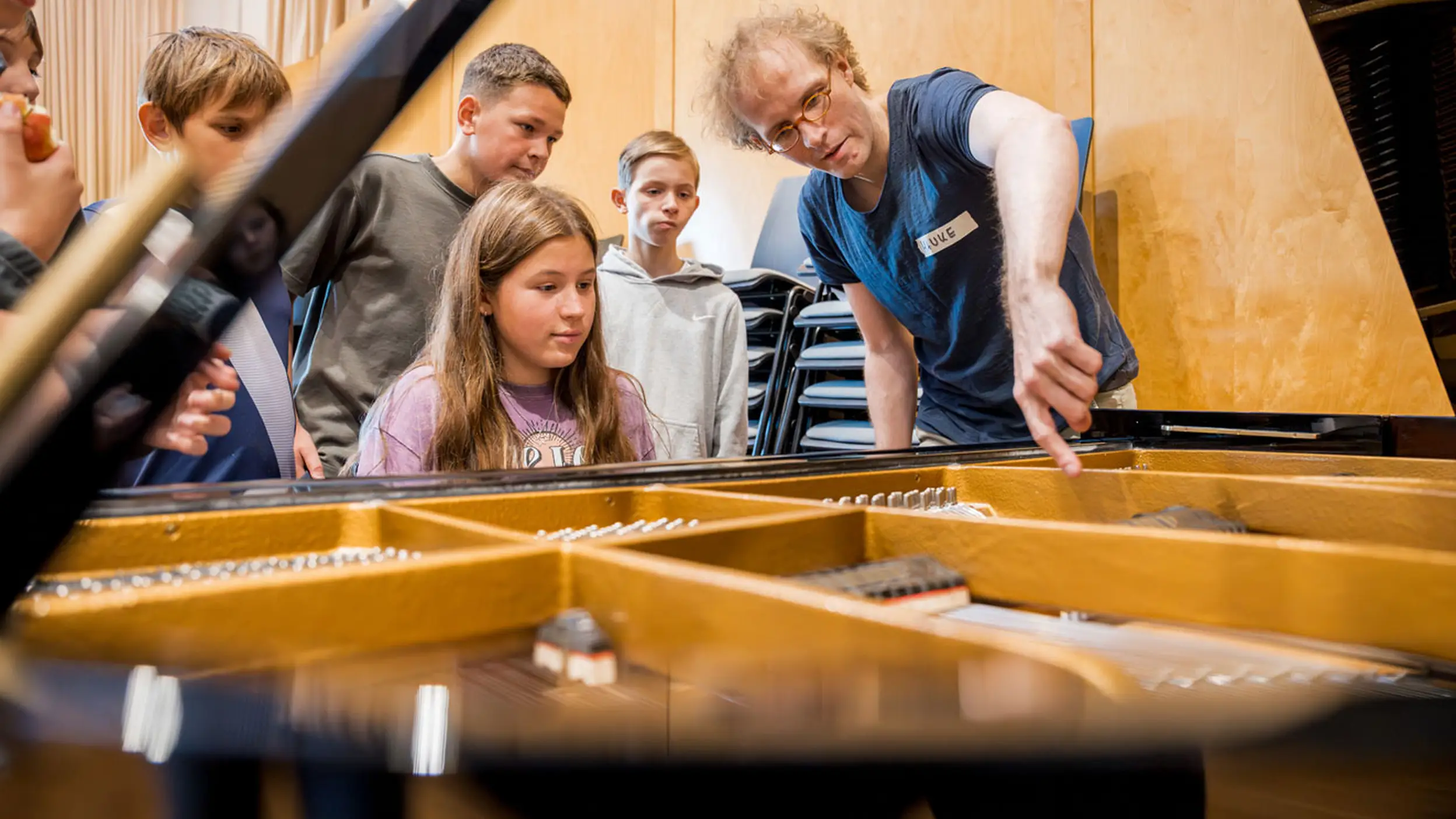 Ein Mädchen sitzt am Klavier und der Lehrer erklärt ihr etwas während die anderen Kinder drumherum stehen und zuschauen.