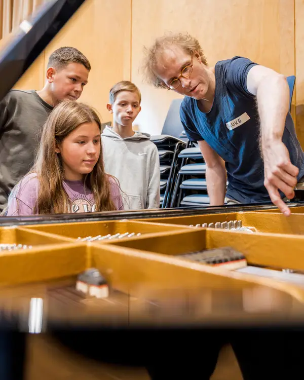 Ein Mädchen sitzt am Klavier und der Lehrer erklärt ihr etwas während die anderen Kinder drumherum stehen und zuschauen.