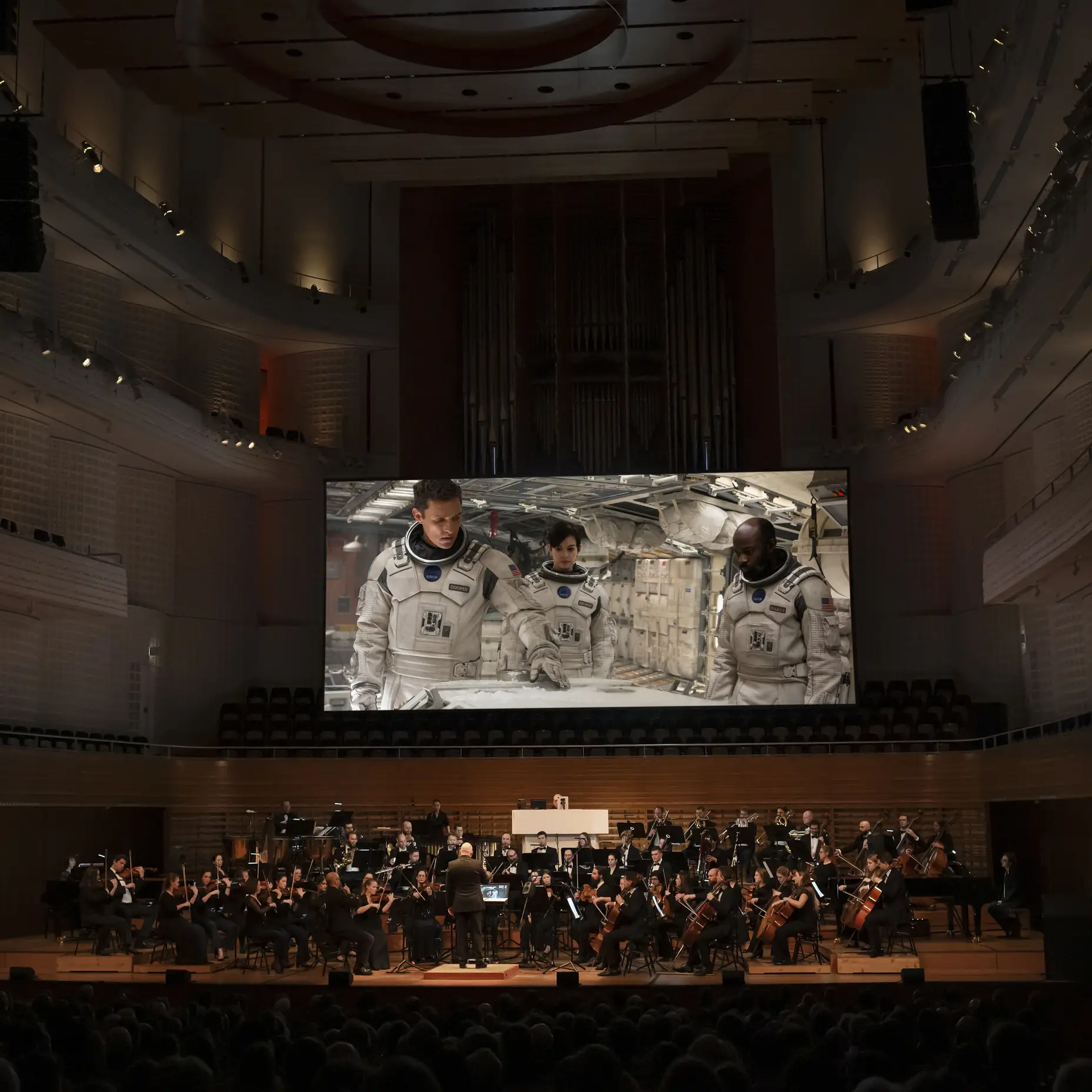 Ein Orchester spielt in einem Konzertsaal. Eine große Leinwand zeigt einen Astronauten im Weltraum. Die Atmosphäre ist eindrucksvoll.