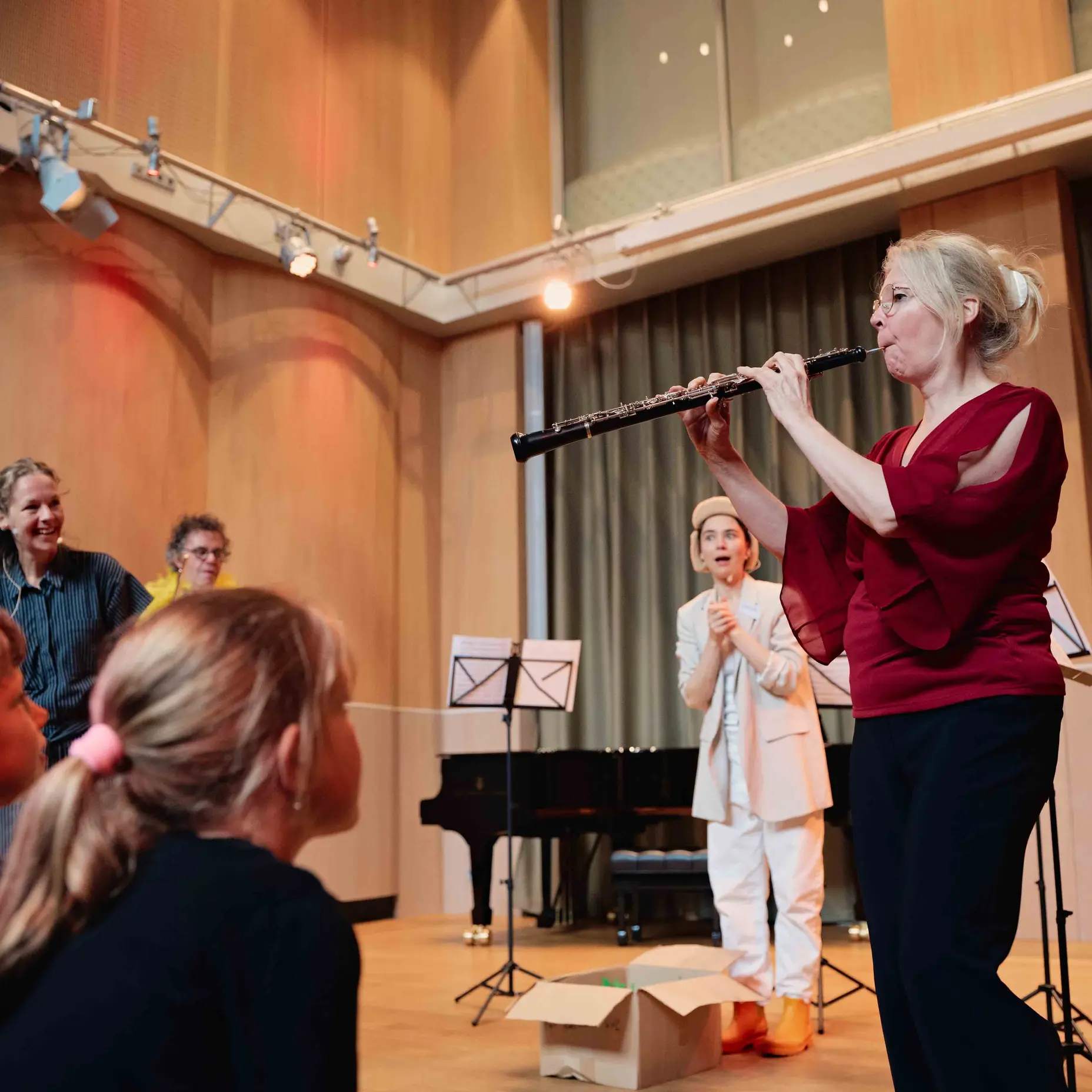 Eine Frau spielt Flöte vor Kindern in einer Aula mit Klavier. Die Kinder sitzen aufmerksam auf dem Boden.