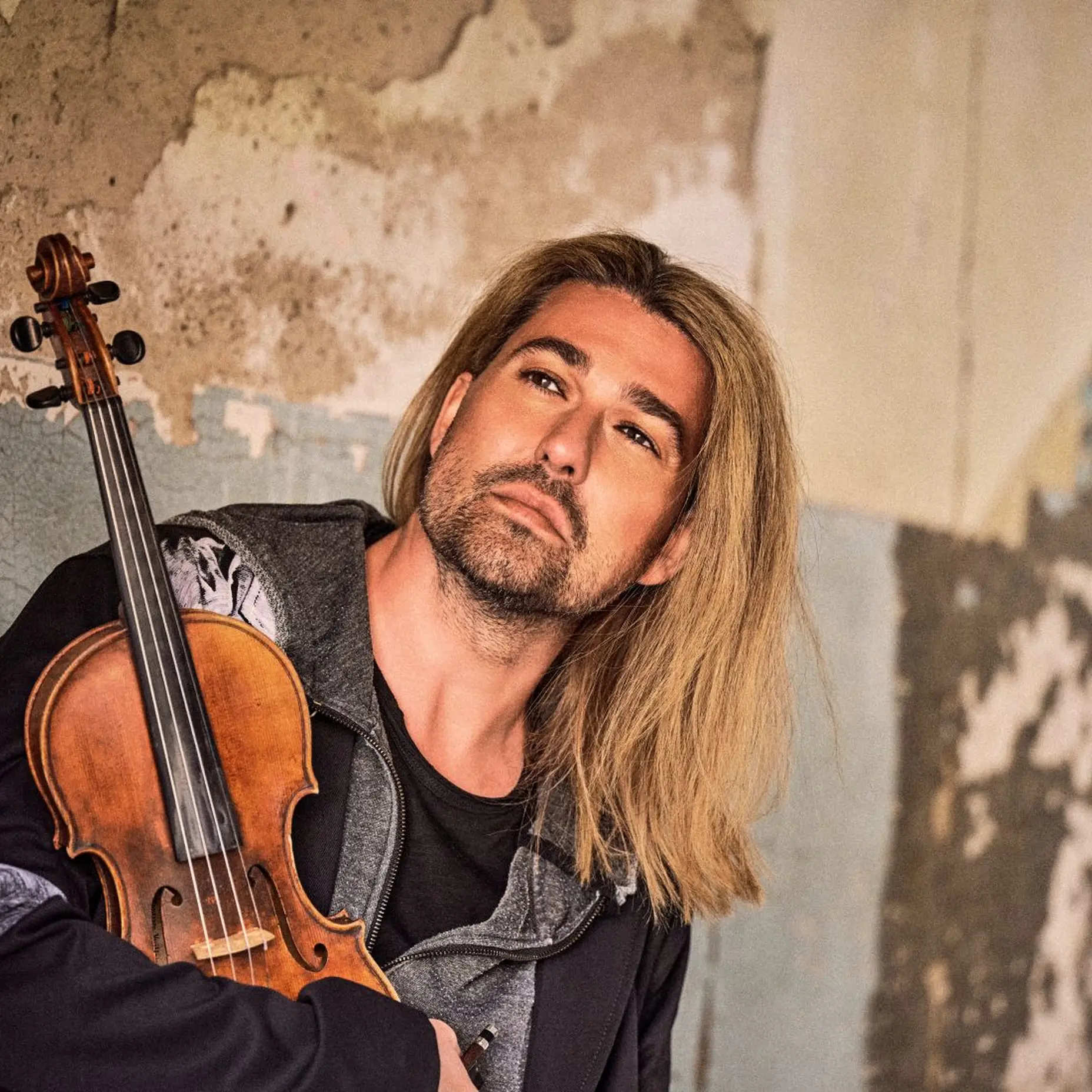 Ein Mann mit langen blonden Haaren hält seine Geige und schaut nach vorne. Er sitzt vor einer Steinmauer und trägt ein grauer Oberteil.