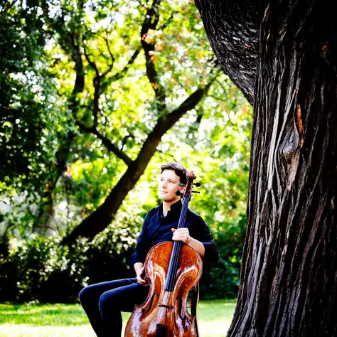 Ein Mann mit einem Cello sitzt auf einer Wiese im Grünen. Er trägt ein dunkelblaues Hemd und Jeans.