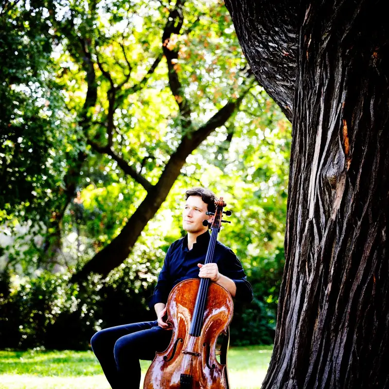 Ein Mann mit einem Cello sitzt auf einer Wiese im Grünen. Er trägt ein dunkelblaues Hemd und Jeans.