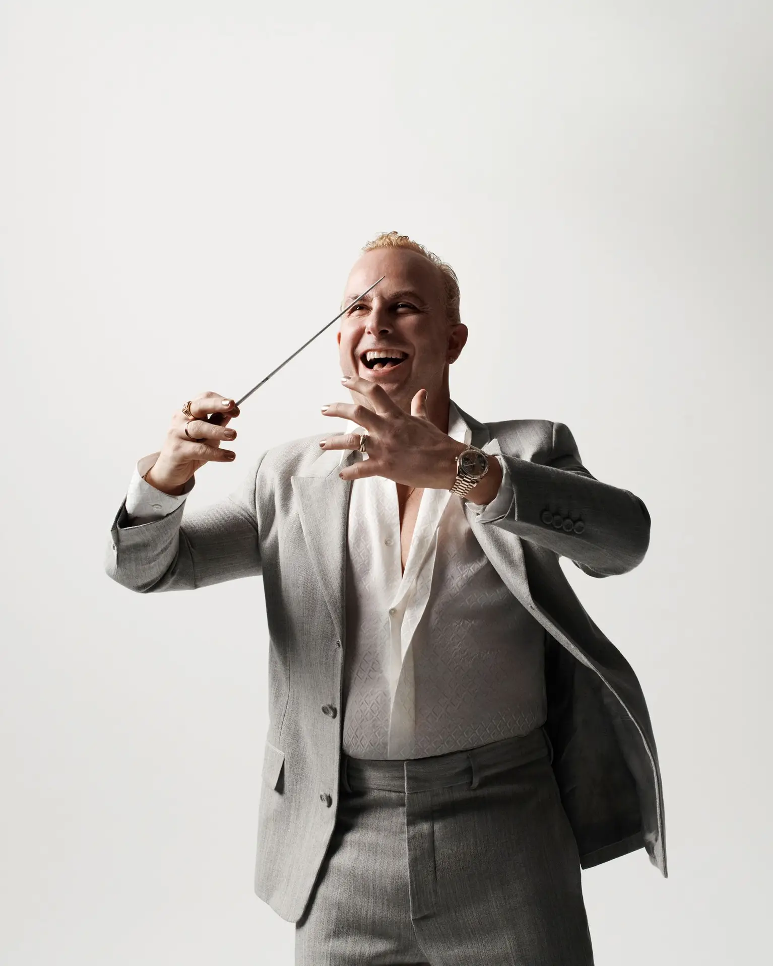 Ein blonder Mann mittleren Alters trägt einen grauen Anzug und ein weißes Hemd. Er lächelt und schwingt seinen Dirigierstab.