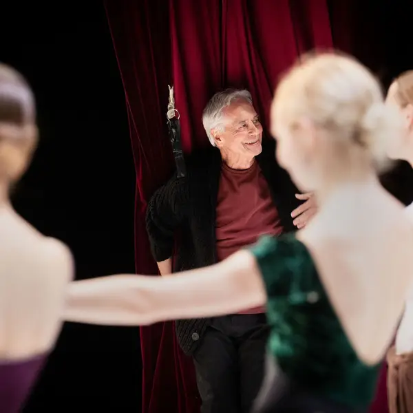 Rechts unscharf im Vordergrund sieht man Balletttänzer und Balletttänzerinnen. Links im Hintergrund sieht man im Fokus einen älteren Mann, der lächelt und ein rotes Shirt trägt.