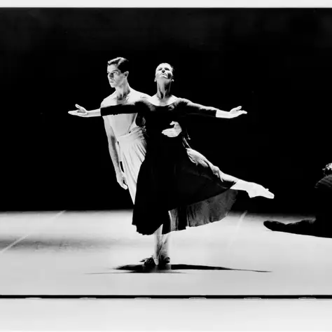 Ein Ballettpaar während einer Aufführung. Die Balletttänzerin trägt ein dunkles Ballettkleid und breitet die Arme auseinander. Der Balletttänzer hinter ihr trägt eine helle Hose und hebt sie.