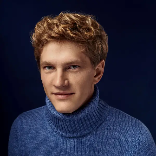 Ein junger Mann trägt einen blauen Rollkragenpullover und sitzt vor einem dunkelblauen Hintergrund. Der Mann hat blonde Haare und lächelt.