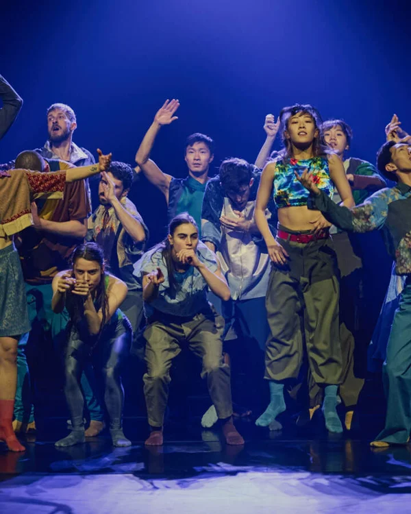 Eine Gruppe von Tänzern auf einer Bühne, in energetischen Posen unter blauem Licht, zeigt moderne Tanzbewegungen.