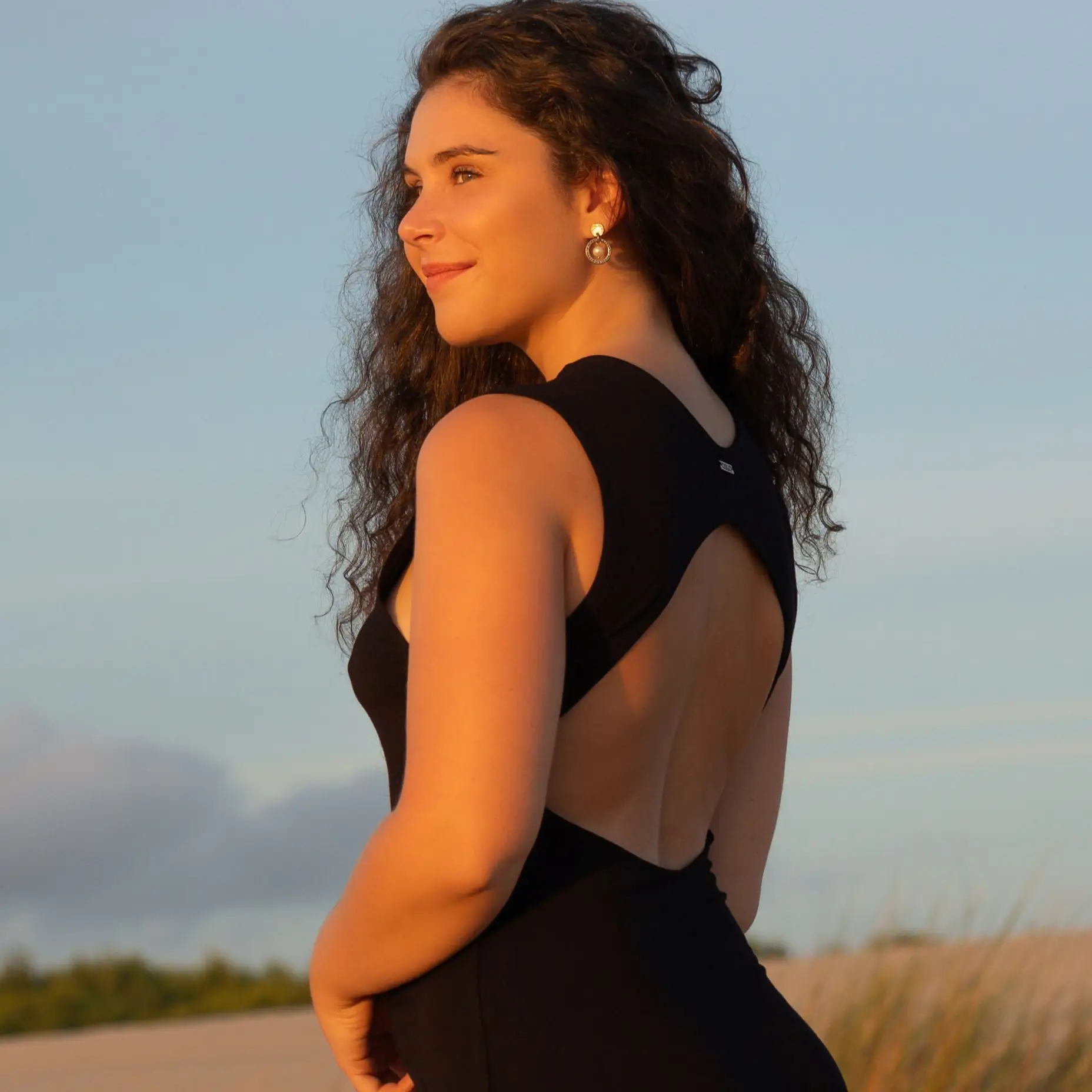 Ein junges Mädchen trägt ihre gelockten braunen Haare offen und wird von der Sonne angestrahlt. Sie trägt ein schwarzes Kleid und steht in der Natur.