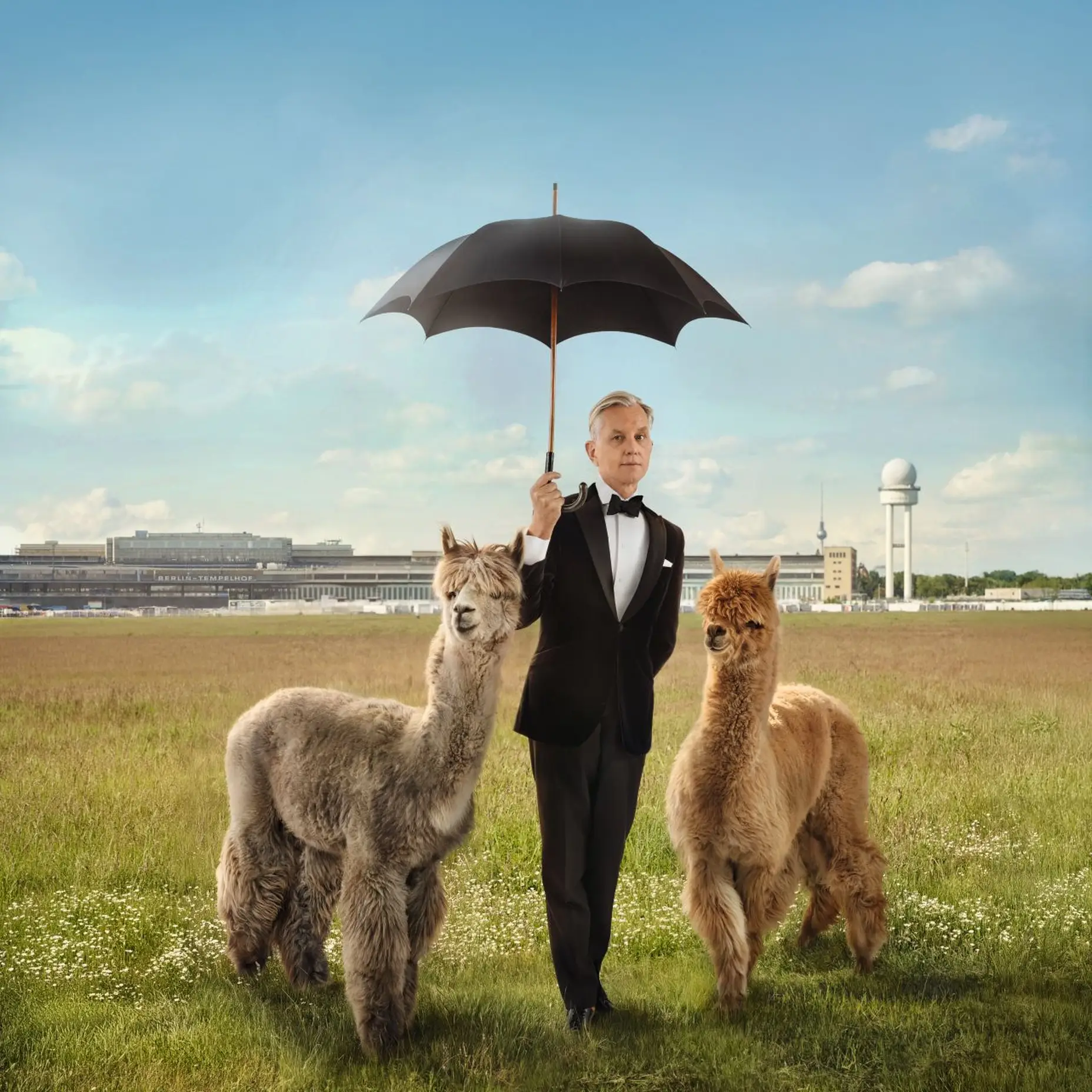 Man sieht einen Mann im Anzug auf einer Wiese und er hält einen Regenschirm. Neben ihm stehen zwei Lamas. Im Hintergrund sieht man den Bahnhof „Berlin Tempelhof".