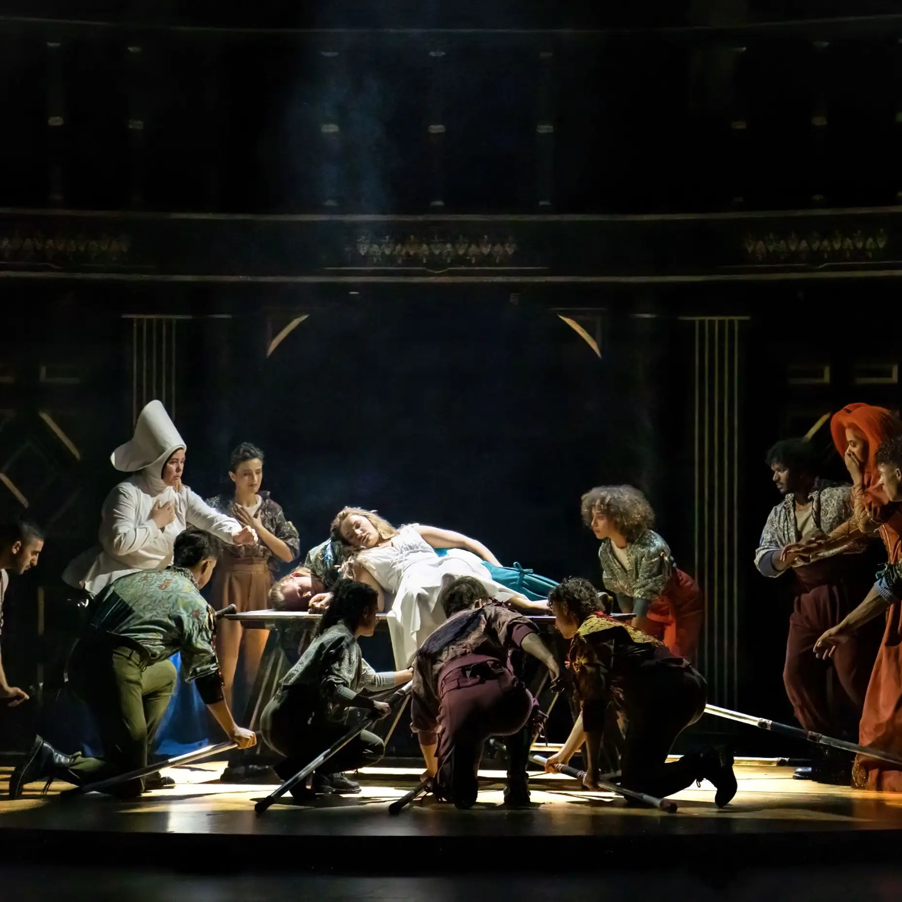 A woman in a white dress and a man are lying on a board on the stage. Many actors are standing around them, looking worried.