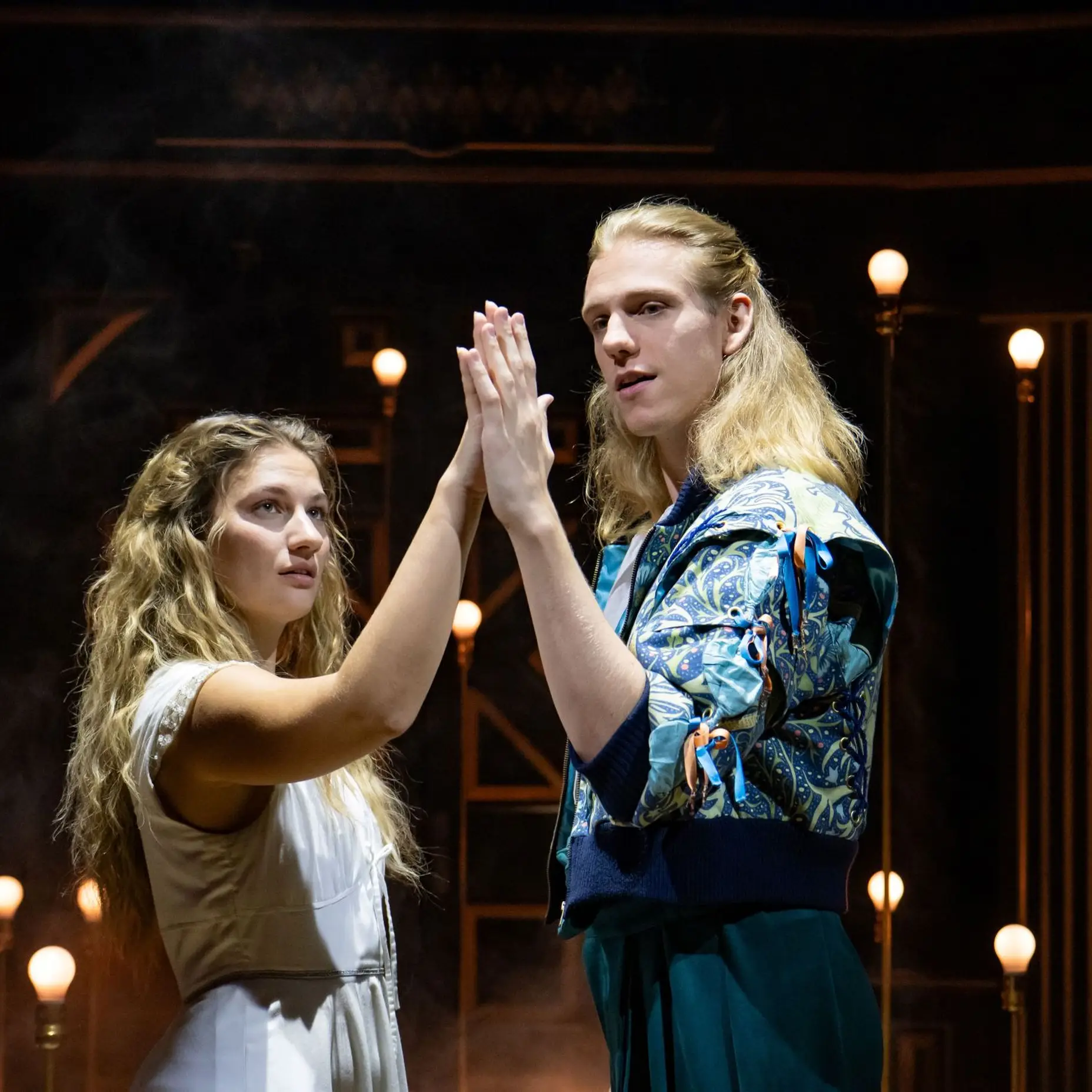 A woman in a white dress with her hair down is standing on the left side of the picture. On the right side of the picture is a man with long blond hair and a patterned jacket. They are holding hands and raising them high; both are looking at their hands.