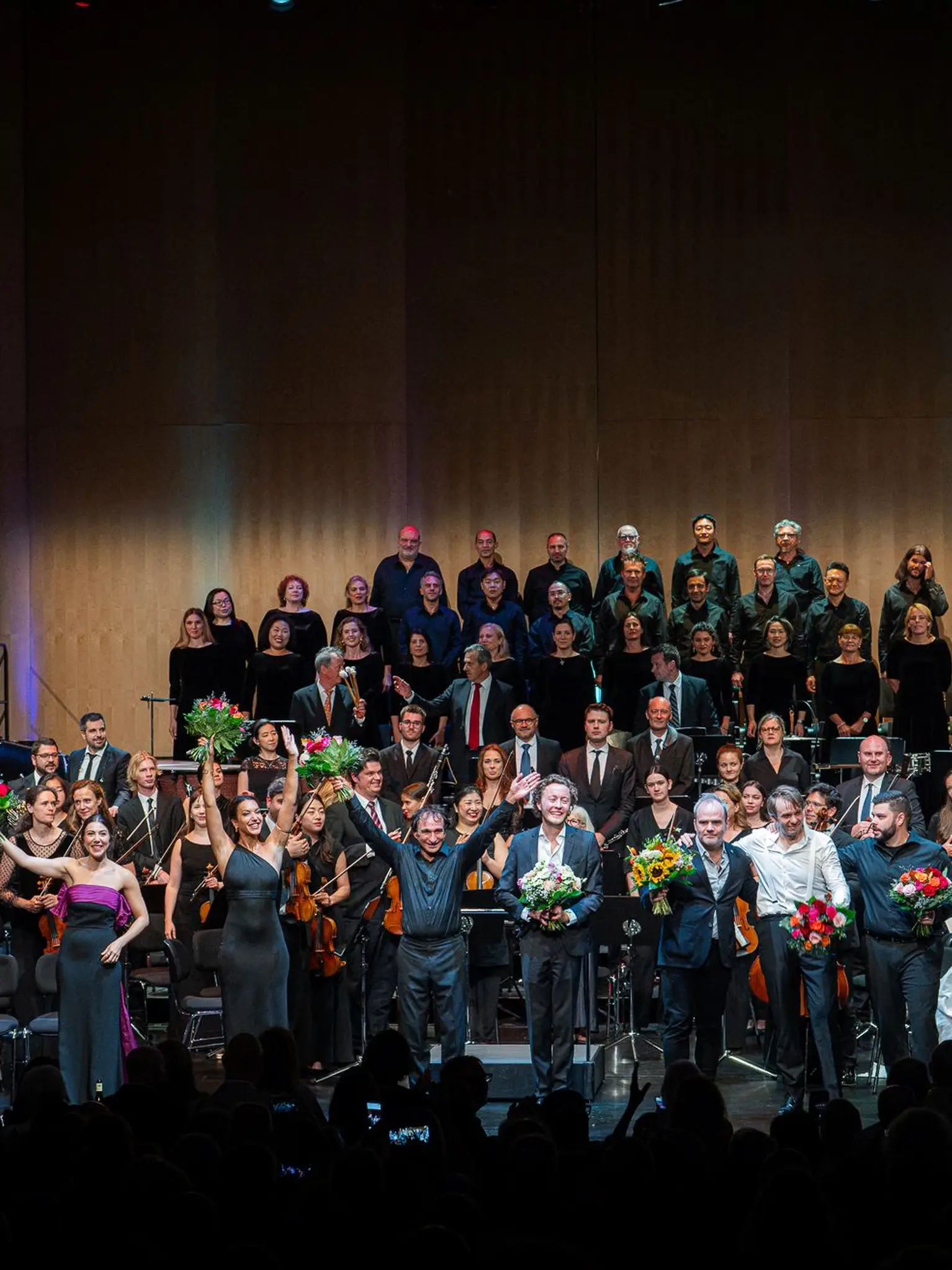 Auf der Bühne sieht man eine Reihe von Opernsängerinnen und Opernsänger zusammen mit dem Dirigenten, die Blumen erhalten haben und sich beim Publikum bedanken. Im Hintergrund sieht man das Orchester und den Chor.