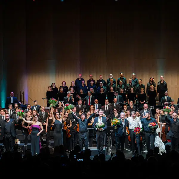 Auf der Bühne sieht man eine Reihe von Opernsängerinnen und Opernsänger zusammen mit dem Dirigenten, die Blumen erhalten haben und sich beim Publikum bedanken. Im Hintergrund sieht man das Orchester und den Chor.