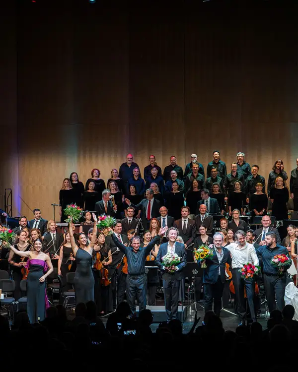 Auf der Bühne sieht man eine Reihe von Opernsängerinnen und Opernsänger zusammen mit dem Dirigenten, die Blumen erhalten haben und sich beim Publikum bedanken. Im Hintergrund sieht man das Orchester und den Chor.