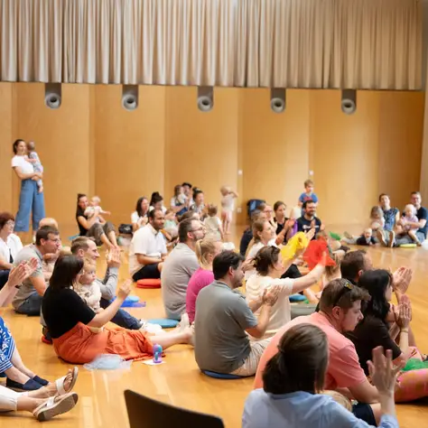 In einem großen offenen Raum sitzen viele Eltern mit ihren Babys und Kleinkindern auf dem Boden und hören Musikern beim spielen der Instrumente zu. Einige klatschen.