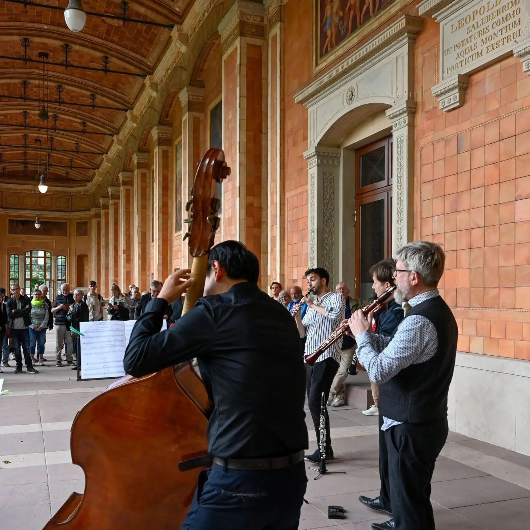 In einem offenen Gebäude sieht man im Vordergrund einen Musiker Kontrabass spielen. Neben ihm spielen andere Musiker Klarinette und Geige. Im Hintergrund sieht man Zuschauer.