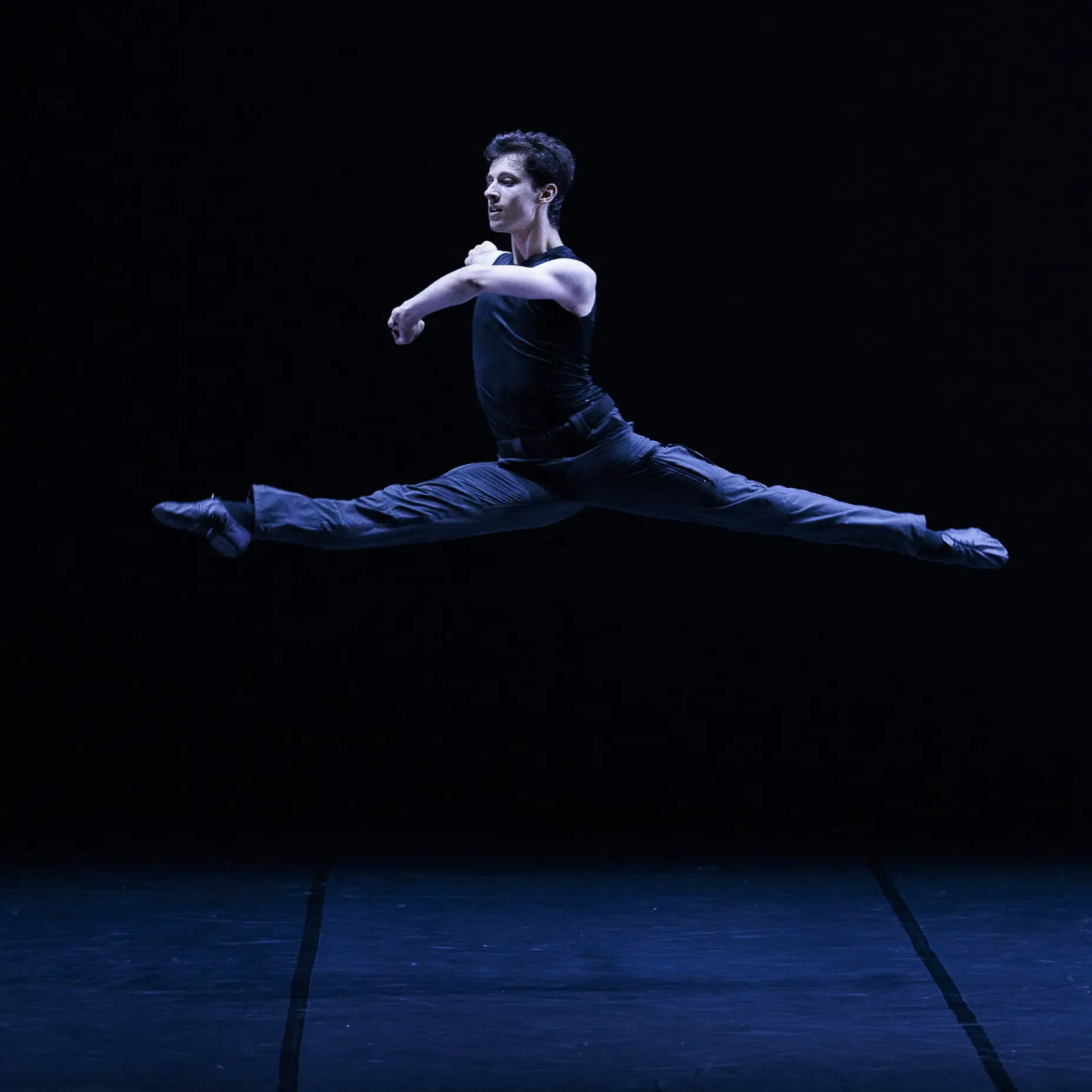 Sur scène, un danseur effectue un grand écart en l'air et lève les bras. Il est vêtu de noir et le fond est noir.